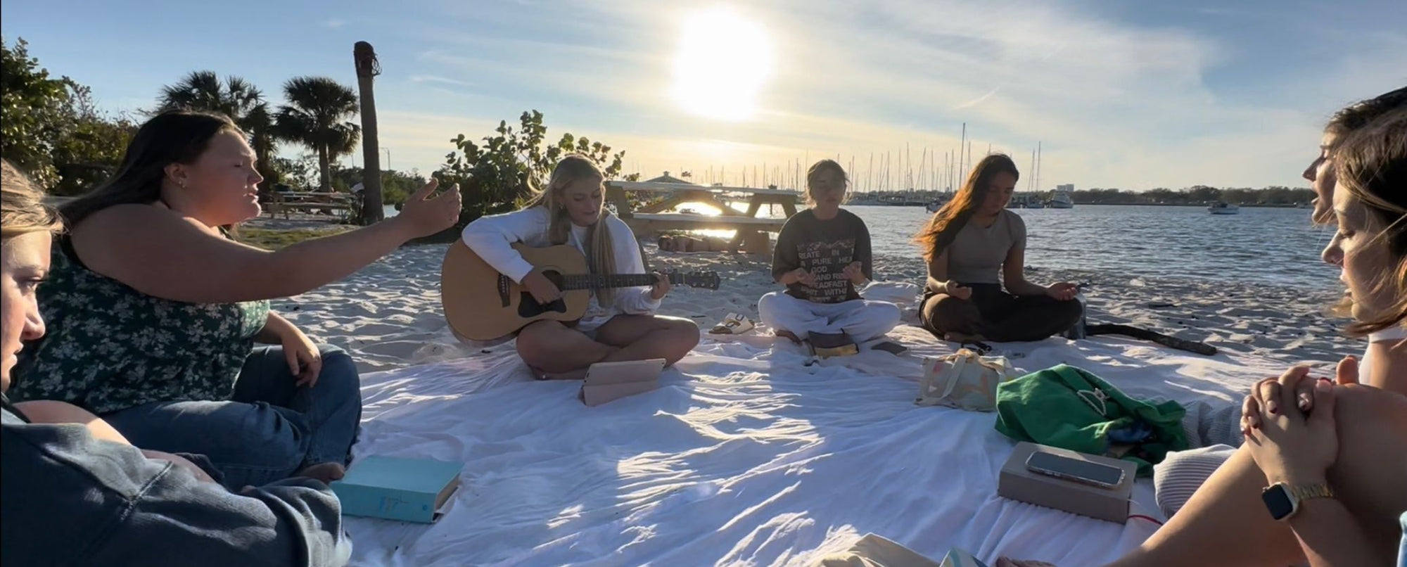 Women relaxing on picnic blanket by the water with guitar, outdoor community gathering at sunset