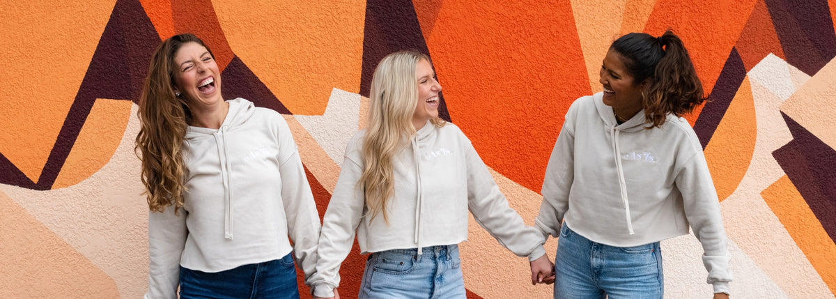 Three women laughing and holding hands in matching neutral hoodies and jeans, standing in front of a colorful mural – casual friendship and lifestyle fashion by As-Is-Sis.