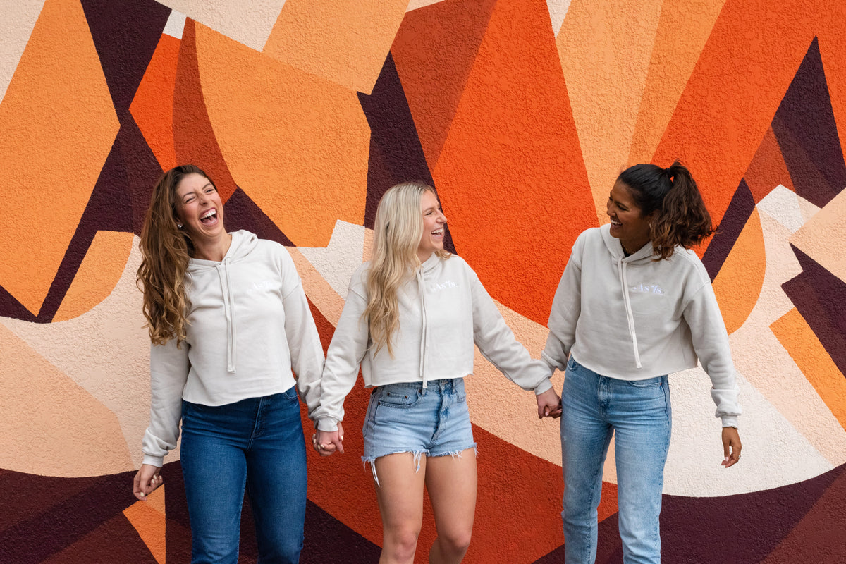 Three women laughing and holding hands in matching neutral hoodies and jeans, standing in front of a colorful mural – casual friendship and lifestyle fashion by As-Is-Sis.